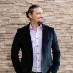 Matthew Young standing in front of a stone wall in a collard shirt and jacket